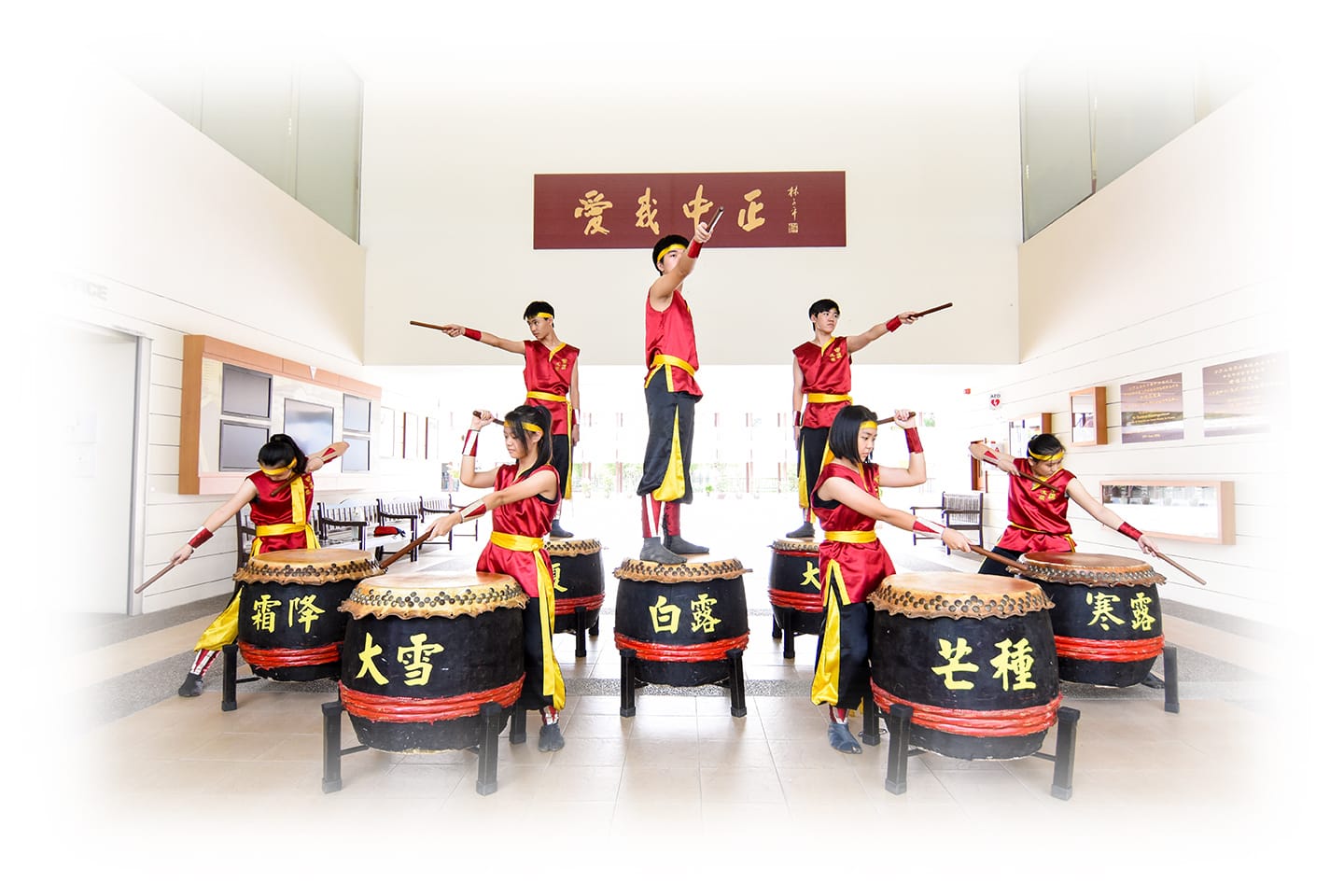 Seven performers in red and yellow costumes play large Chinese drums with sticks indoors, one standing atop a drum.